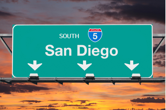 A green highway sign that says in white lettering "South Route 5 - San Diego" with three arrows pointing downwards set against an orange sunset.