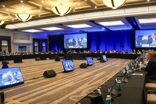 A large conference room with attendees seated around a U‑shaped table, floor monitors and two large screens showing the speaker at the front.